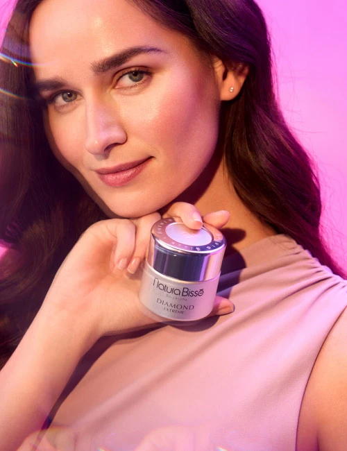 Woman with a slight smile holding Natura Bissé's Diamond Extreme Cream moisturizer with a rich texture. Skincare product featured in bestsellers.
