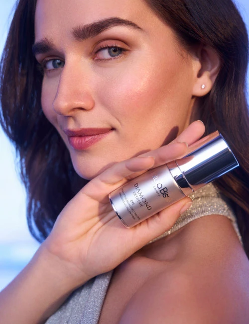 Woman with a slight smile, holding Natura Bissé's Diamond Extreme Eye cream. Skincare product featured in bestsellers.