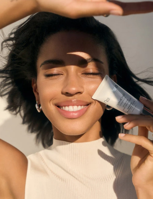 Woman smiling and holding Natura Bissé's Diamond Luminous SPF Sunscreen certified by the Skin Cancer Foundation. Skincare product featured in bestsellers.
